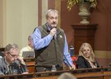 Representative Hansen stands at his desk in the Chamber and speaks into a microphone to someone out of view.